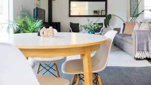 Un salon moderne avec une table ronde en bois, des chaises blanches et un canapé gris. On aperçoit des plantes et un téléviseur à l’arrière-plan.