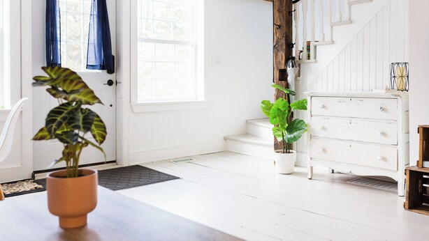 Una habitación luminosa con una escalera de madera, un aparador blanco y dos plantas en macetas.