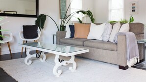 Uma sala de estar com uma mesa de centro de vidro, um sofá cinza e uma poltrona branca.