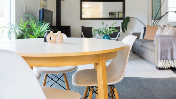 Uma sala de estar moderna com uma mesa de madeira redonda, cadeiras brancas e um sofá cinzento. Há plantas e uma televisão ao fundo.