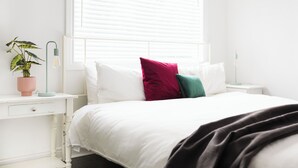 Cama bem arrumada com roupa de cama branca e várias almofadas, mesa de cabeceira com abajur e planta em vaso no criado-mudo.