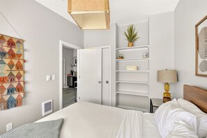 A hotel room with a bed, a bedside table, a lamp, a shelf with decorative items, and a door leading to a hallway.