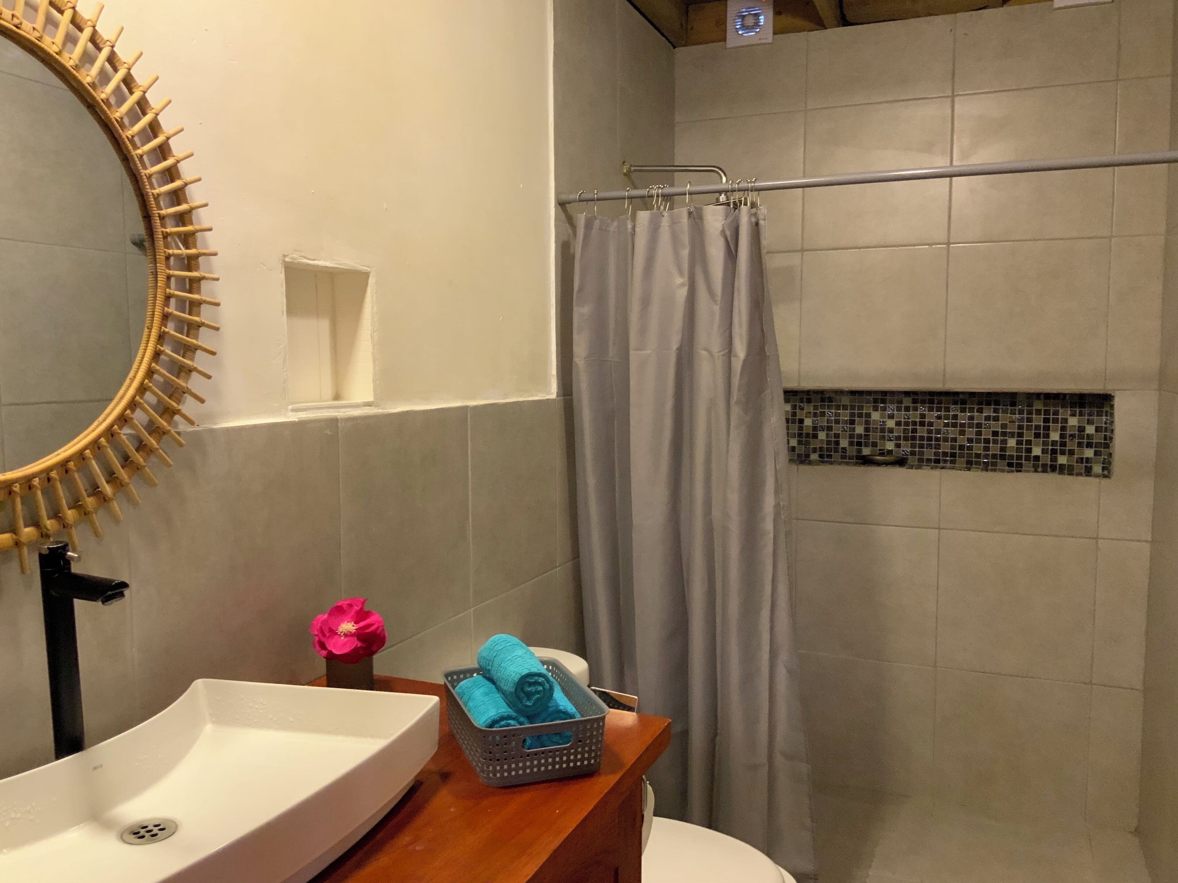 Une salle de bain avec un lavabo blanc, un meuble-lavabo en bois, un rideau de douche et une petite étagère fixée au mur, ornée d’une fleur rose.