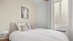 A bedroom with a bed, a bedside table, a lamp, a wall-mounted picture, and a window with curtains. - Renovated Apartment Min. from Beach/Palais (Cannes)