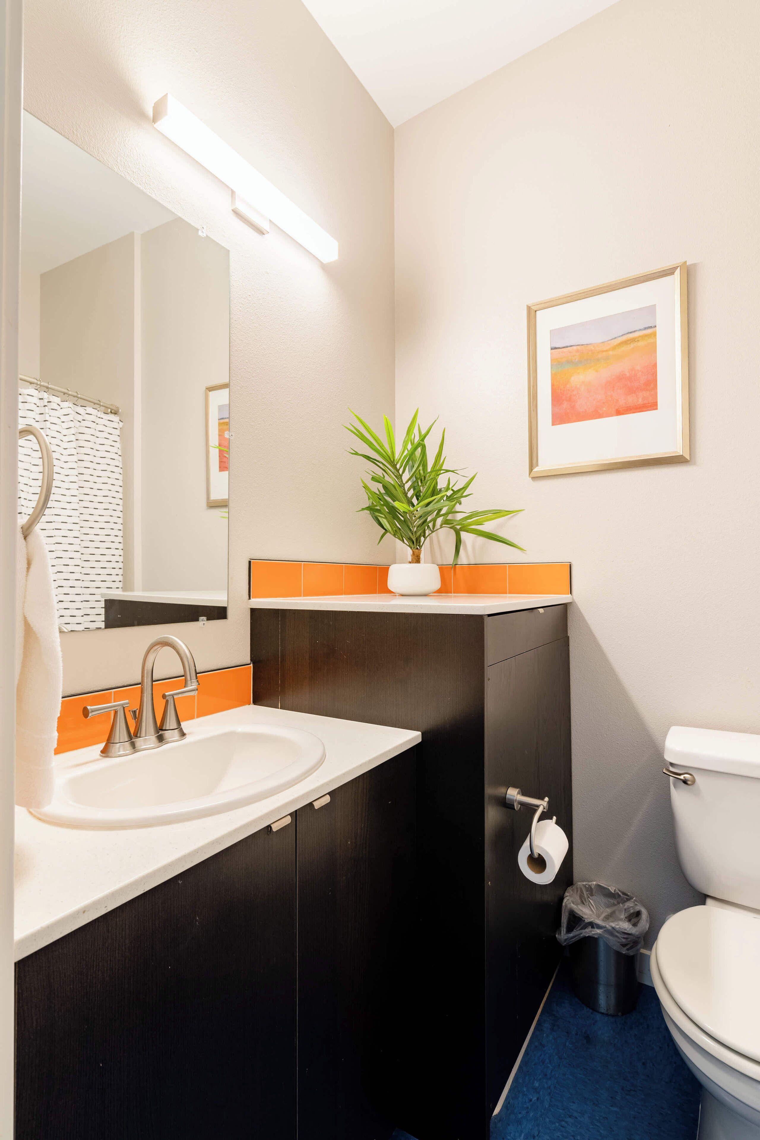 A bathroom with a white sink, black cabinet, and a toilet. There is a plant on the counter and a framed picture on the wall.