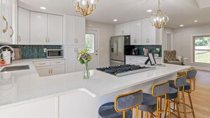 A modern kitchen with white cabinetry, a large island with bar stools, stainless steel appliances, and decorative lighting. - Poolside Paradise-Kayaks/SUPs, Private Pool, Billiards, Foosball (Winchester)