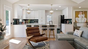 A modern kitchen with white cabinets, a central island, and bar stools. There is a brown leather chair and a grey sofa in the living area. - Poolside Paradise-Kayaks/SUPs, Private Pool, Billiards, Foosball (Winchester)