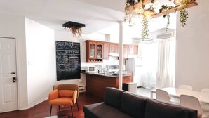 Un salon moderne avec un canapé, une table basse en verre et un fauteuil orange. On peut apercevoir au fond un tableau noir où sont écrits des objectifs et un réfrigérateur.