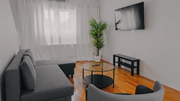 Ein modernes Wohnzimmer mit grauem Sofa, einem runden Glastisch, einem schwarzen Beistelltisch, einer Topfpflanze und einem an der Wand montierten Fernseher.
