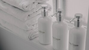Drei weiße Flaschen mit den Aufschriften 'PURE SHAMPOO' und 'PURE BODY WASH' auf einer Fensterbank neben gefalteten weißen Handtüchern.