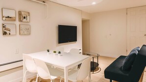 Uma sala de estar moderna com uma mesa branca, cadeiras brancas, um sofá preto e uma TV de tela plana fixada na parede.