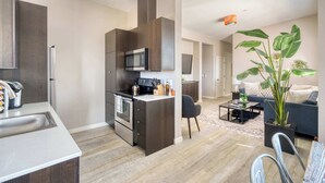 Une cuisine moderne avec des meubles de couleur sombre, un four à micro-ondes et un four encastré. On y trouve un coin repas avec une table et des chaises, ainsi qu’un salon avec un canapé et une grande plante.