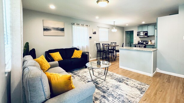A living room with a grey sofa, yellow cushions, a coffee table, and a patterned rug. There is a dining area with chairs and a table, and a kitchen with a countertop and cabinets.