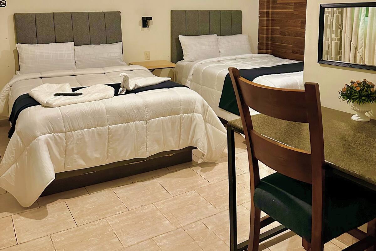 A hotel room with two beds, a wooden headboard, a dining table with a chair, and a small table with a vase of flowers.