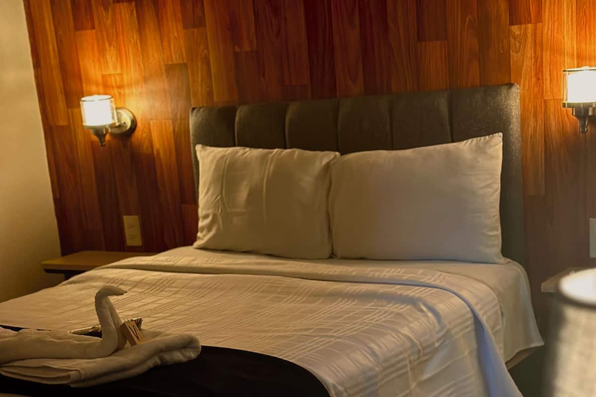 A hotel room with a wooden headboard, a bed with white linens, a nightstand, and a lamp.