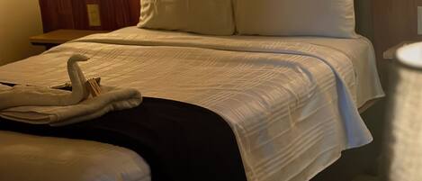 Una habitación de hotel con cabecera de madera, cama con ropa blanca, mesita de noche y lámpara.
