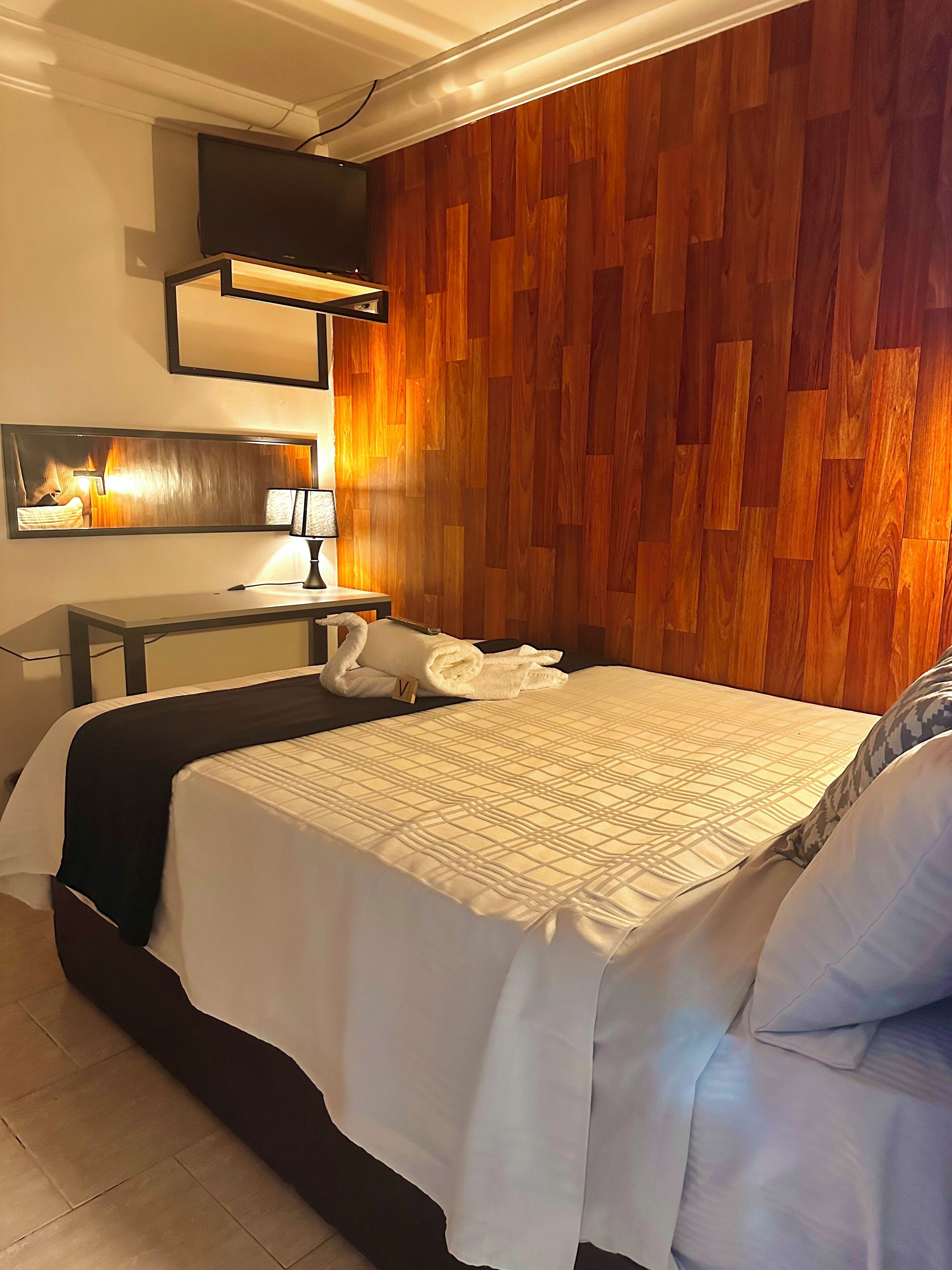 Habitación de hotel con cama, escritorio, silla, televisor de pantalla plana, espejo y panel de pared de madera.