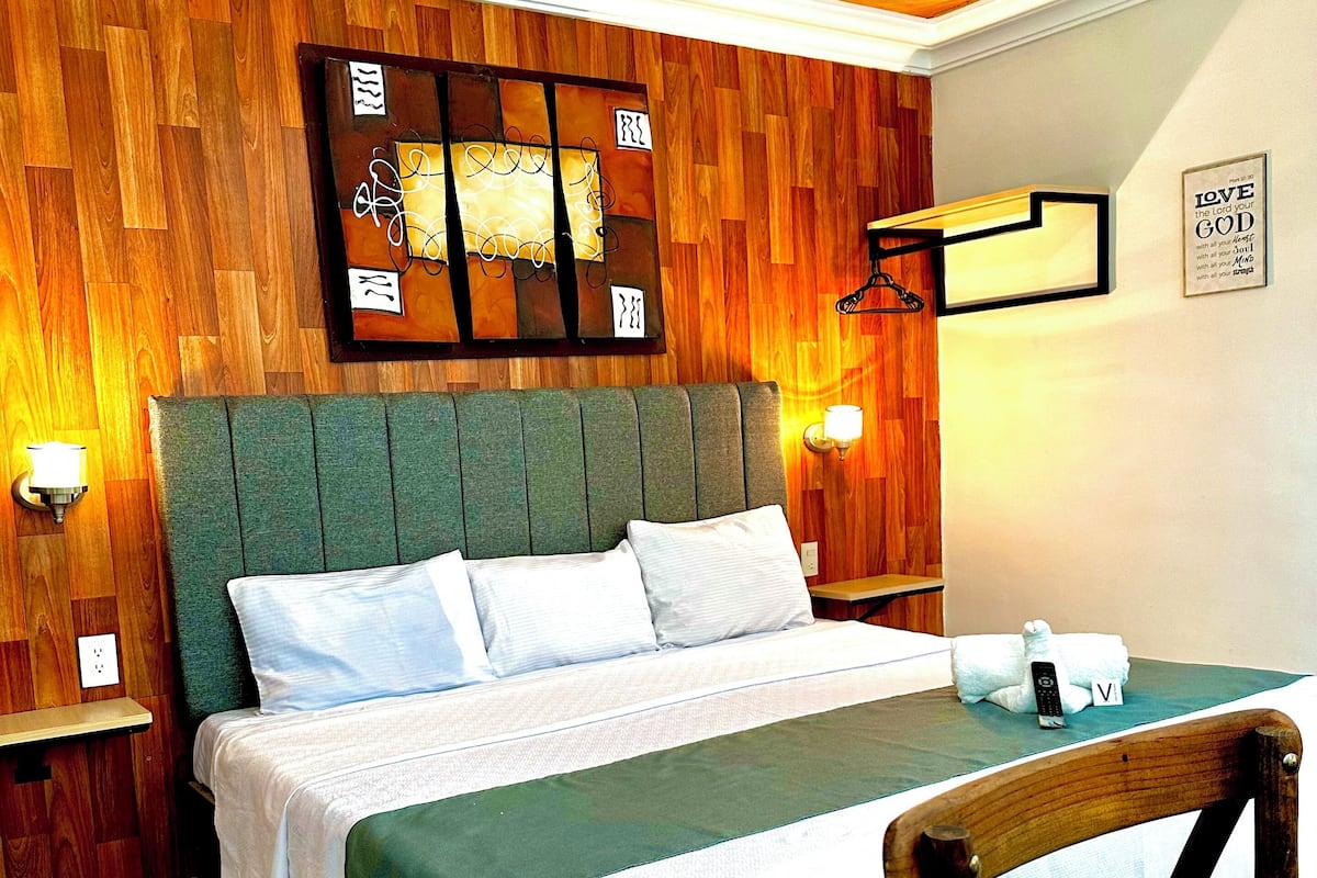 A neatly made bed with a green headboard, a wooden wall panel, and a wall-mounted light fixture.