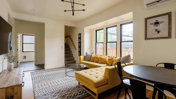 A modern living room with a yellow sofa, a round dining table, and a staircase leading to an upper level. - 1920s Farmhouse | 4BR/2BA | Foodie District (Portland)