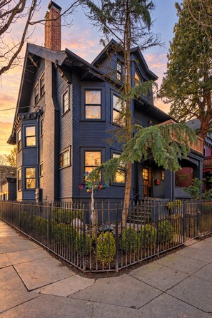A two-story house with a dark exterior, a visible chimney, and a gated front yard. - Historic 2BR | Walk to NW 23rd Shops & Eats (Portland)