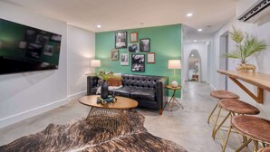 Ein modernes Wohnzimmer mit einem schwarzen Ledersofa, einem runden Couchtisch und einer grünen Akzentwand mit gerahmten Bildern. Ein Fernseher ist an der Wand montiert, ein kleiner Beistelltisch mit Lampe steht daneben, sowie eine Holztheke mit Hockern.
