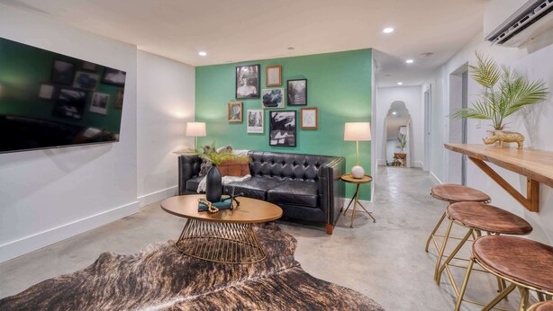 A modern living room with a black leather sofa, a round coffee table, and a green accent wall with framed pictures. There is a TV mounted on the wall, a small side table with a lamp, and a wooden bar with stools. - Historic Nob Hill 2BR | Prime Walkable Spot (Portland)