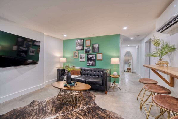 A modern living room with a black leather sofa, a round coffee table, and a green accent wall with framed pictures. There is a TV mounted on the wall, a small side table with a lamp, and a wooden bar with stools. - Historic Nob Hill 2BR | Prime Walkable Spot (Portland)