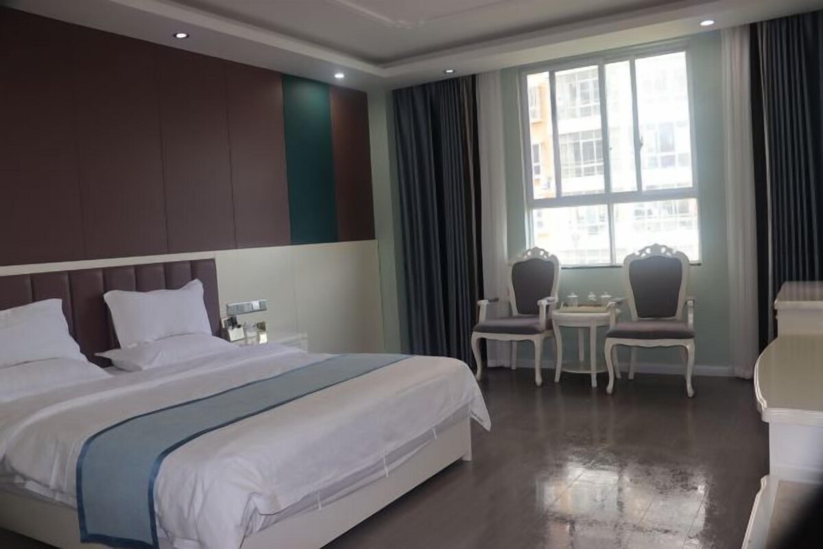 Een hotelkamer met een groot bed, twee nachtkastjes, een stoel, een klein tafeltje en een raam met gordijnen.