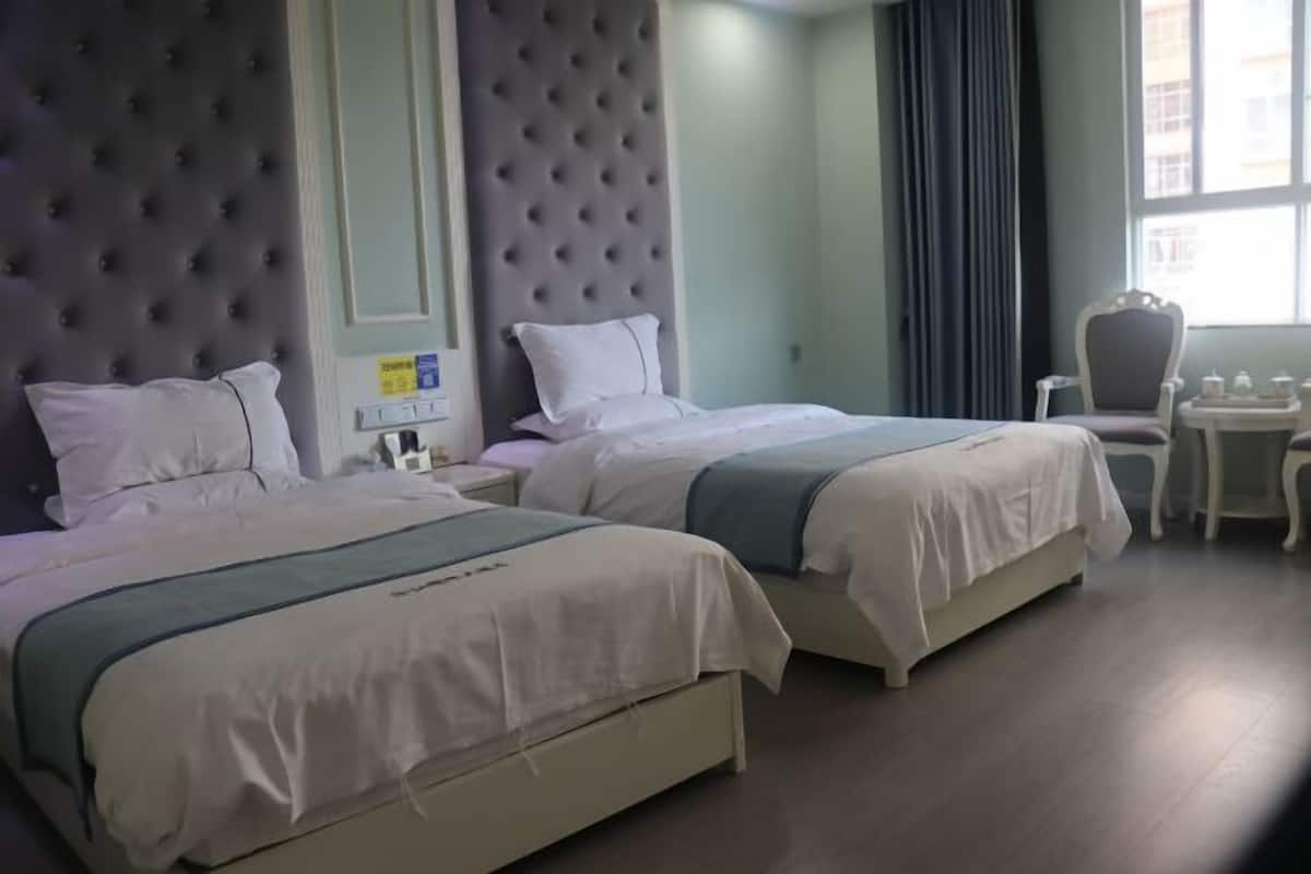 Hotellihuone, jossa on kaksi sänkyä, koristeltu pääty, pieni pöytä ja tuoli sekä ikkuna, jossa on verhot.