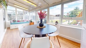 Un coin repas avec une table ronde, des chaises blanches, un vase rempli de fleurs roses et une vue sur le jardin à travers de grandes fenêtres.