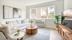 Un salon moderne avec un canapé gris, une table basse ronde en bois, un vase blanc orné de fleurs, un téléviseur à écran plat posé sur un meuble en bois, et une grande fenêtre donnant sur des immeubles.