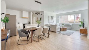 Une cuisine moderne avec des meubles blancs, un coin repas avec une table et des chaises en bois, et un salon avec un canapé et un téléviseur.