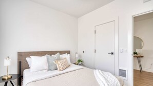 A neatly arranged bedroom with a bed, bedside table, lamp, and a door leading to another room. - Alberta Arts - 2BR - Walk to Food, Shops, & Art (Portland)