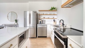 A modern kitchen with a stainless steel refrigerator, dishwasher, and oven. There are wooden shelves above the counter holding dishes and a potted plant. - Sleeps 12 - Exclusive Floor - Great for Groups (Portland)
