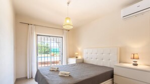 A bedroom with a bed, bedside tables, a hanging light fixture, an air conditioning unit, and a window with curtains.
