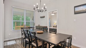 Un coin repas avec une table et des chaises en bois foncé, un lustre et de grandes fenêtres équipées de stores.