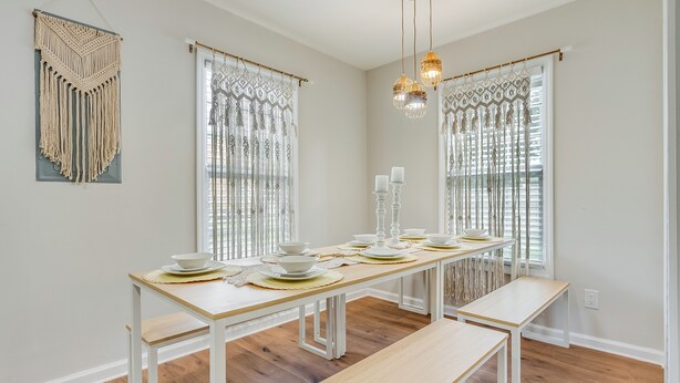 Een eethoek met een lange tafel gedekt voor vier personen, witte wanden, houten vloer en een decoratieve macramé wandversiering.