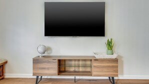 Een moderne woonkamer met een flatscreen televisie aan de muur, een houten console tafel met een wereldbol en een plant, en een glazen plank.