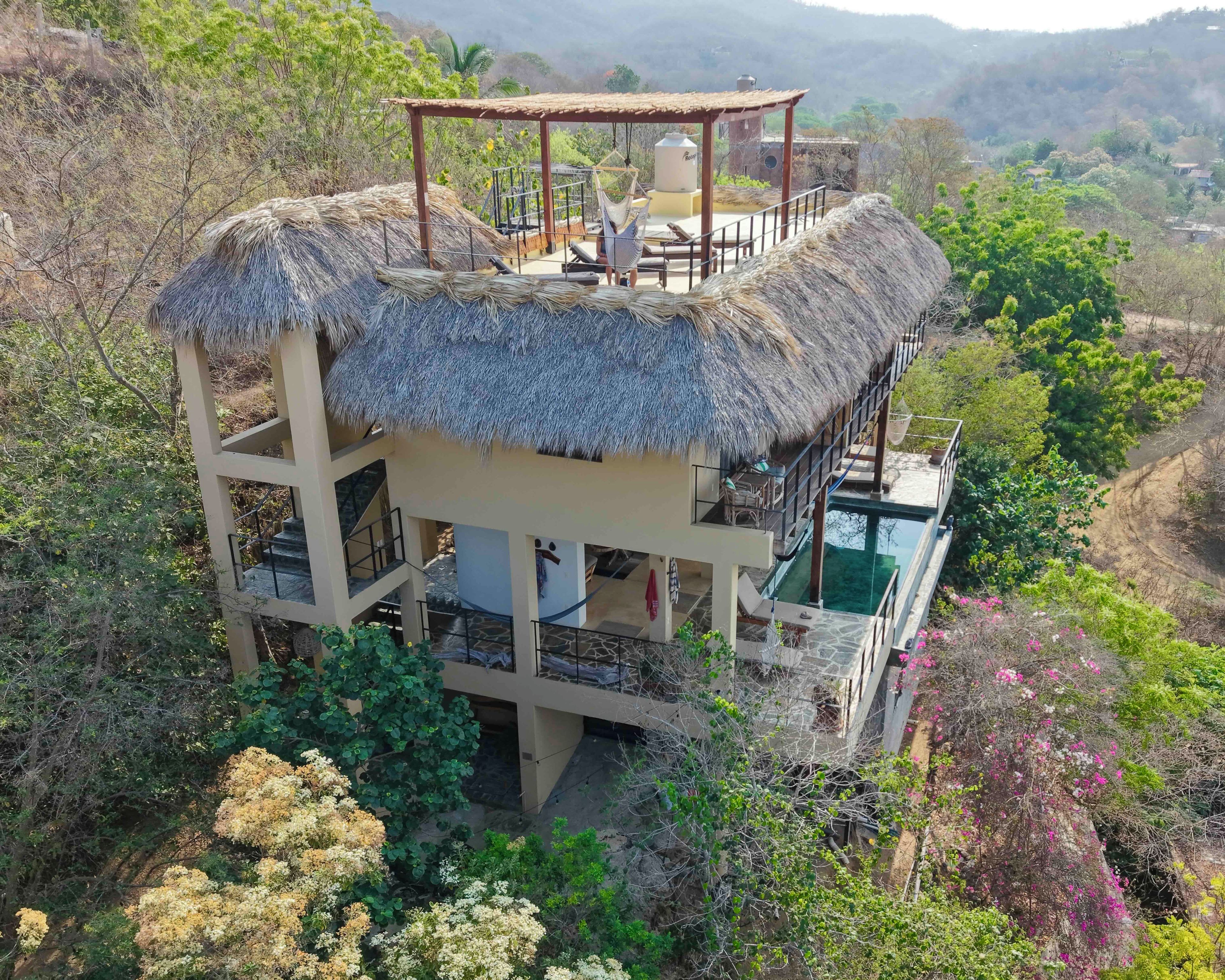 Uma construção com telhado de palha, várias varandas, piscina e vegetação exuberante ao redor.