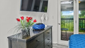 Une chambre avec un téléviseur fixé au mur, une commode avec des tiroirs, un vase rempli de fleurs rouges et un ventilateur.