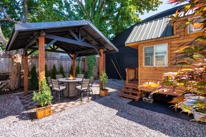 A covered patio with a fire pit, outdoor seating, and a gravel ground. - The Sunstone • Modern Tiny House • Prime Location (Portland)