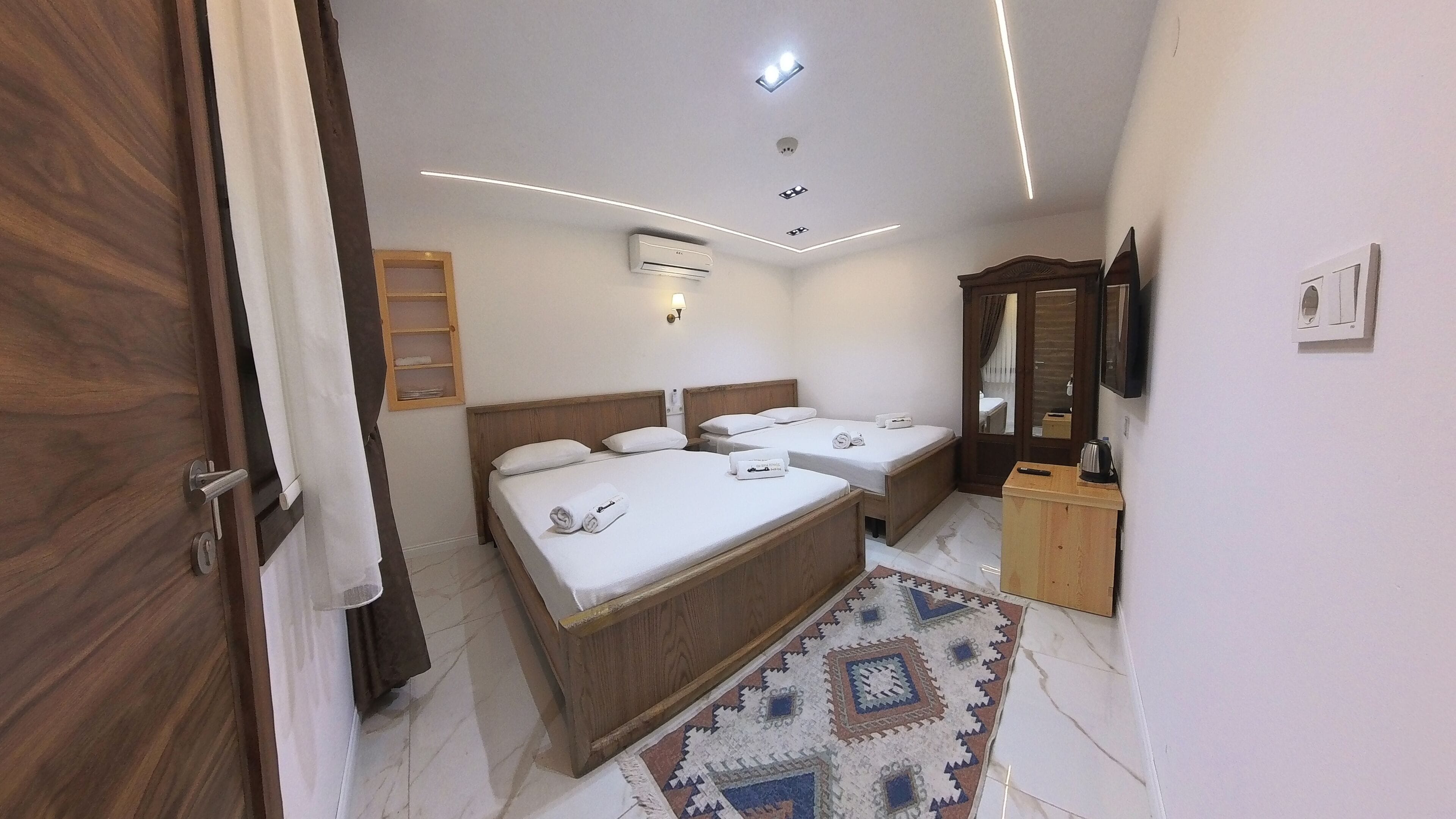 A hotel room with two beds, a wooden wardrobe, a small cabinet, a mirror, and a ceiling-mounted air conditioner.