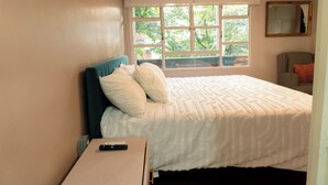 Una cama bien tendida con edredón blanco y almohadas, una mesita de noche con control remoto y una ventana que da a vistas de árboles y un edificio.