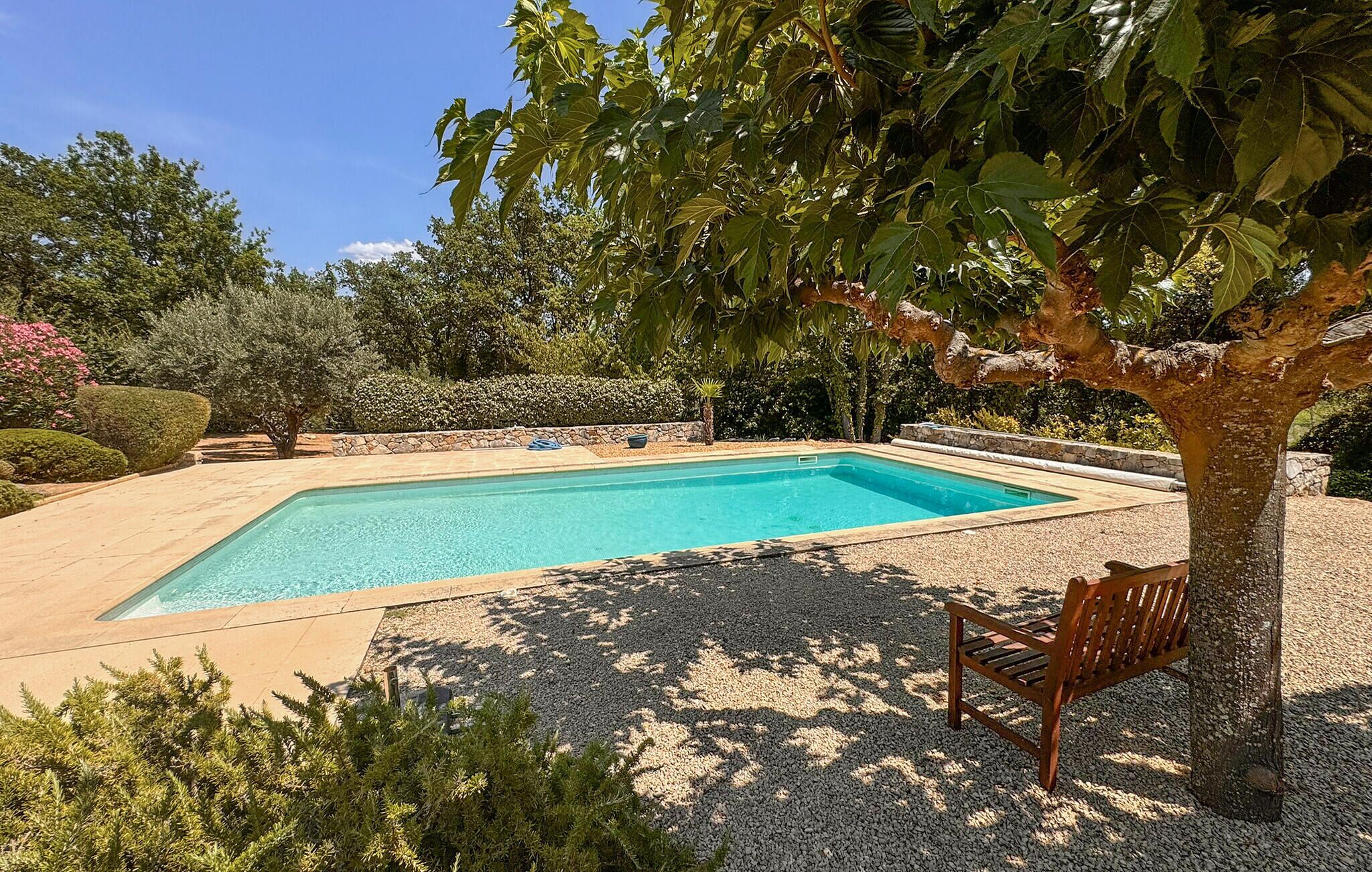 Une piscine rectangulaire bordée de galets, entourée d’un jardin aménagé avec différents arbustes et un banc.