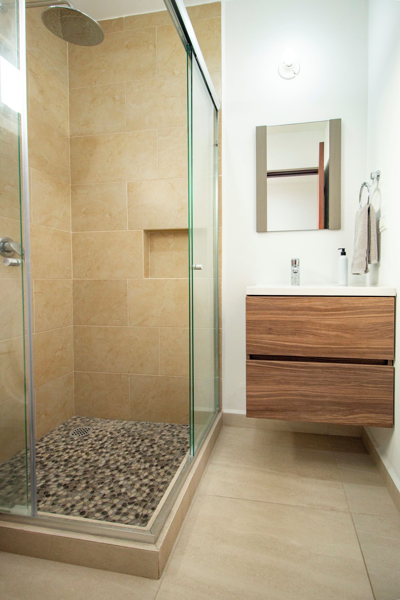 Un baño moderno con ducha de vidrio, azulejos color beige, un mueble de tocador de madera y un espejo arriba.