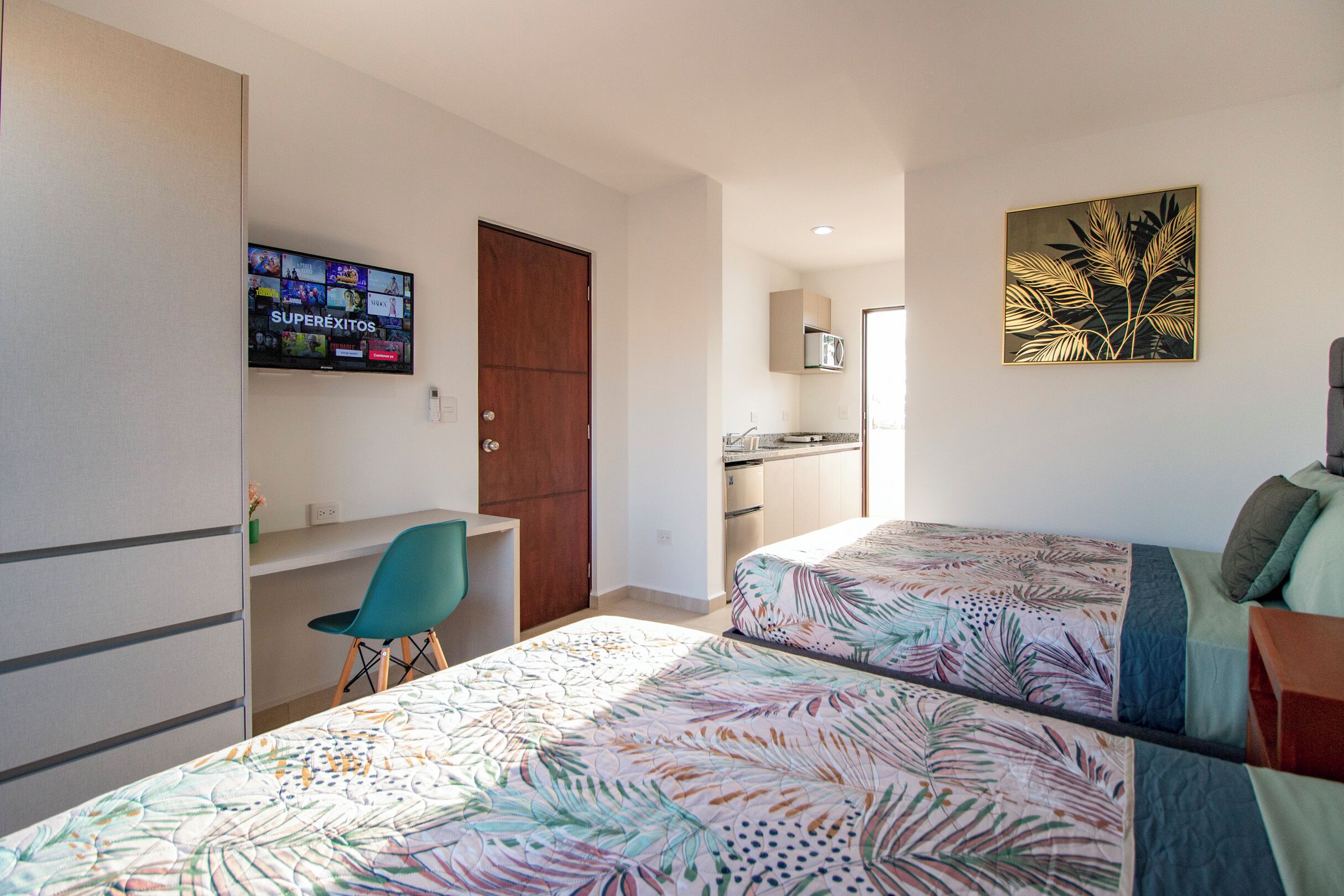 A bedroom with two beds, a desk, a chair, a TV, and a kitchen area.