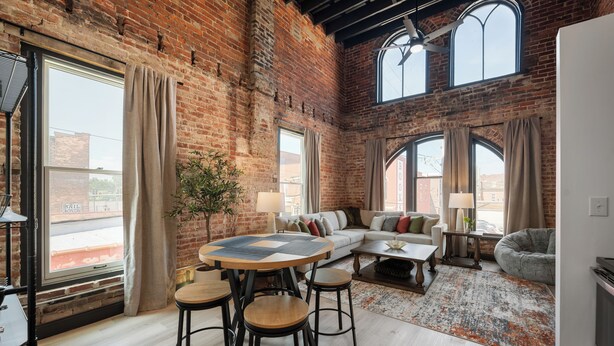 A spacious loft with exposed brick walls, large windows, a round wooden table with stools, a sofa, and a bean bag. - Cozy 1-bedroom apartment in fabulous Wabash with WiFi, AC (Wabash)