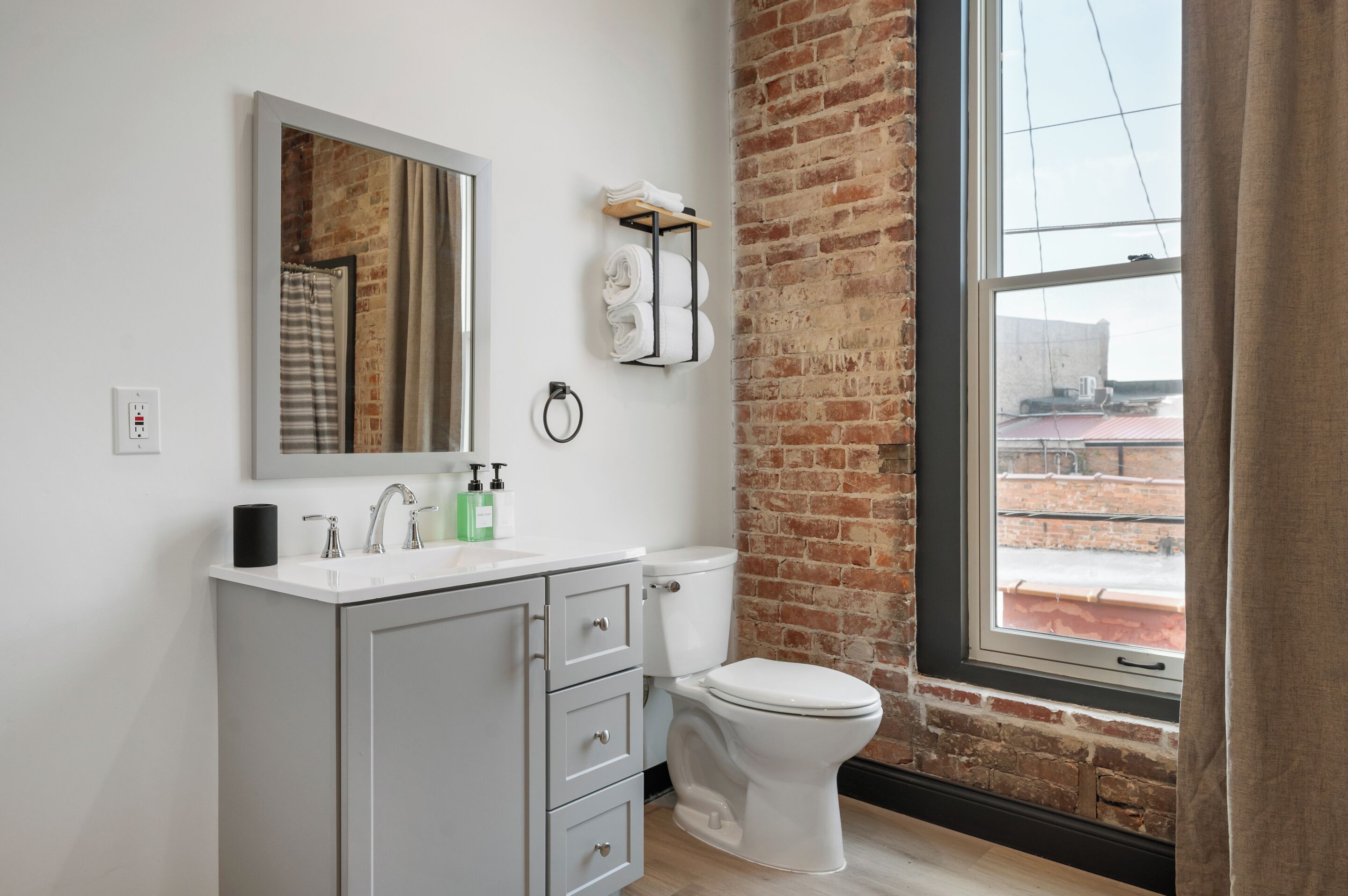 A modern bathroom with a white sink, mirror, and toilet. It features a brick wall and a large window with a view of the outside.