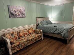 Une chambre avec un canapé à motifs floraux, un lit recouvert d’une couverture grise et un tableau mural.
