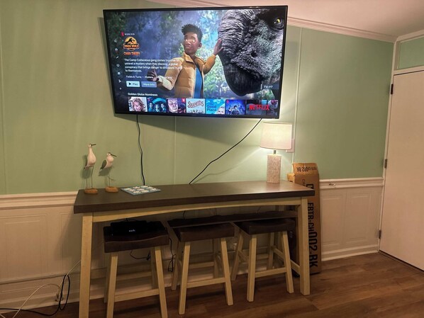 Un salon avec un téléviseur à écran plat fixé au mur, une table basse en bois, deux tabourets de bar et une lampe.